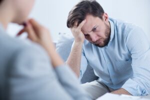 An upset man in a blue shirt listens to his therapist as he holds his head in his hand.