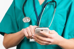 A nurse wearing teal scrubs and a stethoscope touches her phone's screen.